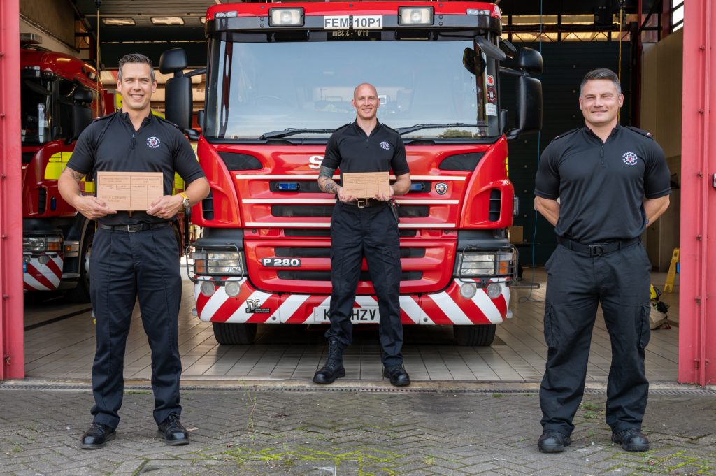 Firefighters Dean Keeber, Aaron Childs and Paul Webb standing in front of a fire engine with trophies