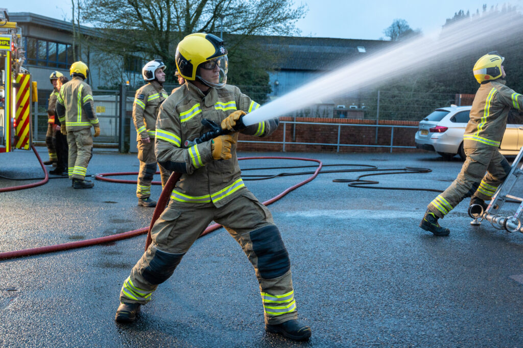 Firefighter using hose to squirt water whilst other firefighters complete ladder drill in the background