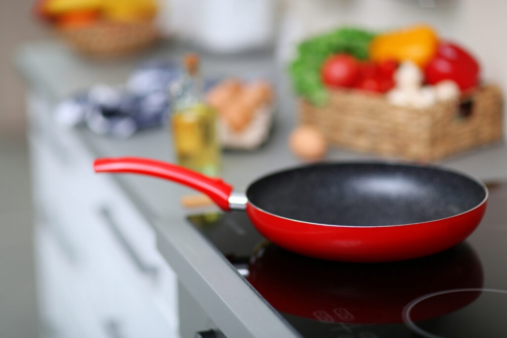 A red frying pan is shown on a hot kitchen hob, with a number of food items which are out of focus behind it