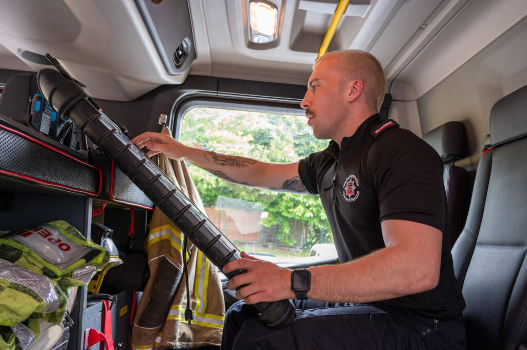 Daventry firefighter Kieran Davies, wearing his black shirt uniform, checks that an item of equipment is working on the back seat of the fire engine.