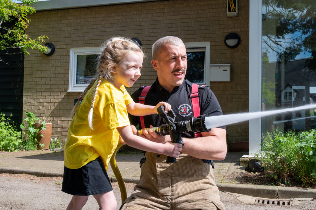 A young schoolgirl wearing a yellow school uniform is shown how to use a hose reel jet by Daventry firefighter Kieran Davies, who is squirting water from the hose.