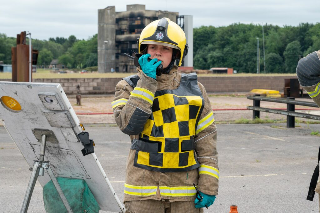 A firefighter in tunic and helmet speaks with a colleague over the radio.
