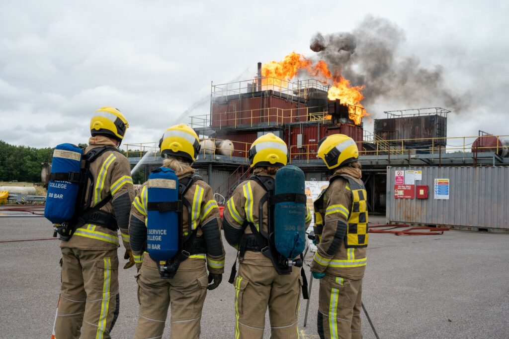 Four firefighters are shown in the forefront, while in the background a flame comes out of a mock oil rig at the Fire Service College.