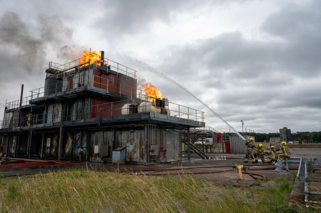 A giant mock oil rig is on fire, with firefighters on the right hand side tackling the blaze.
