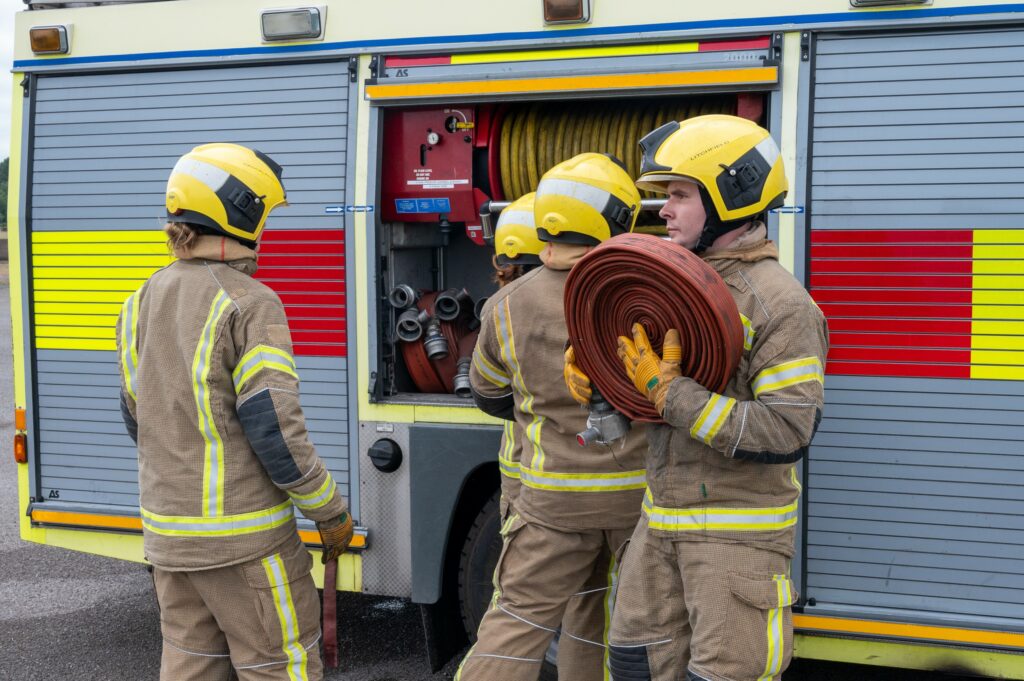 Three firefighters are shown in front of a fire engine, with the one on the right holding a red hose.