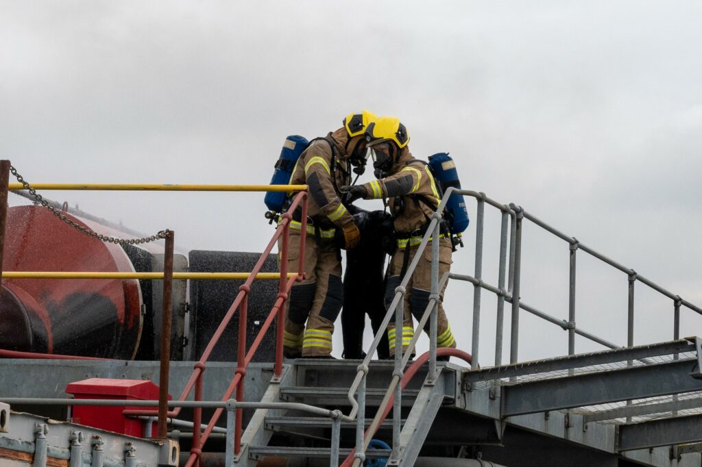Two firefighters wearing yellow helmets and fire tunic are shown dragging a dummy down a flight of stairs during a training exercise.