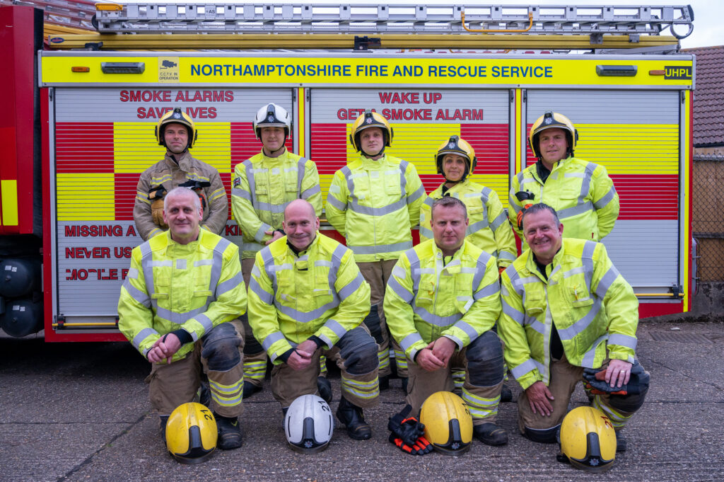 9 firefighters stood by the side of a fire engine