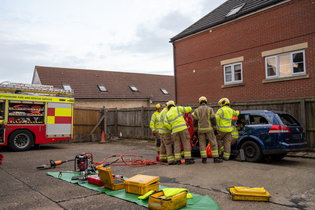 4 firefighters cutting up a car with a fire engine in the background