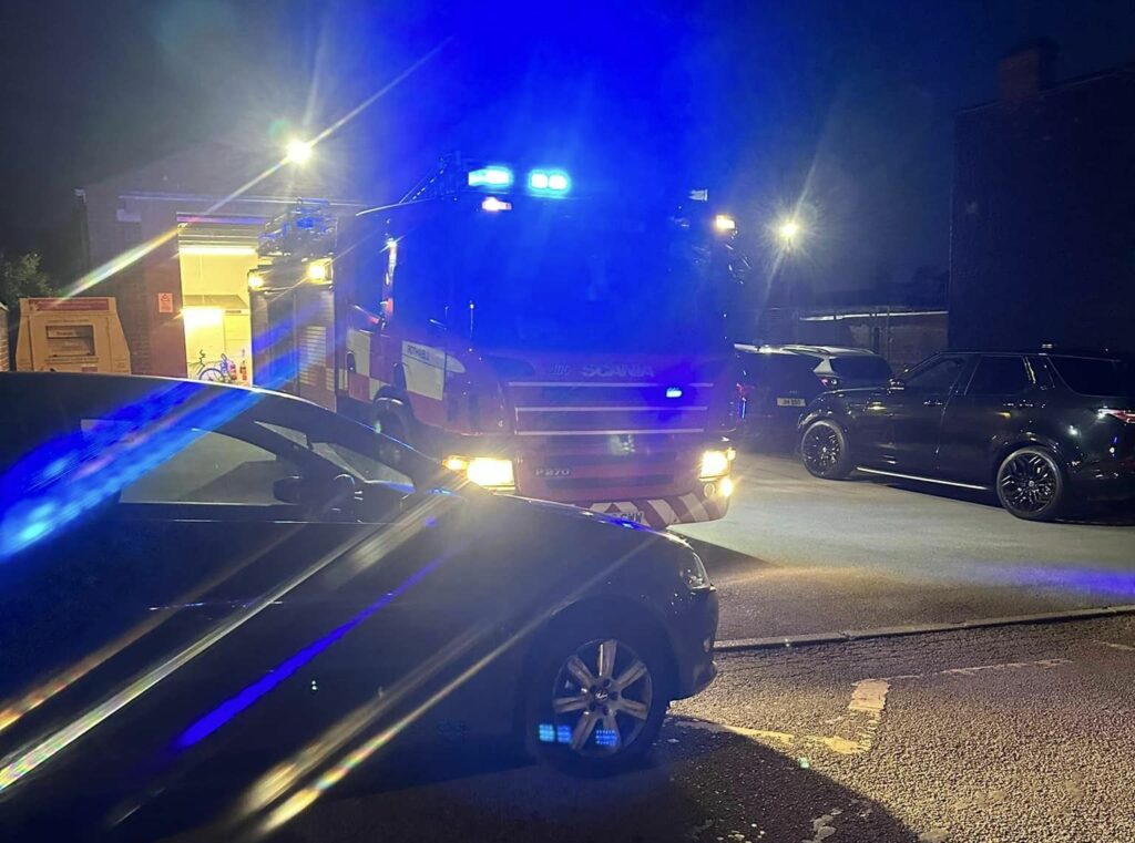 A black car on the left is shown partially blocking the Rothwell fire engine - with its flashing blue lights - as it attempts to leave the forecourt at the Station