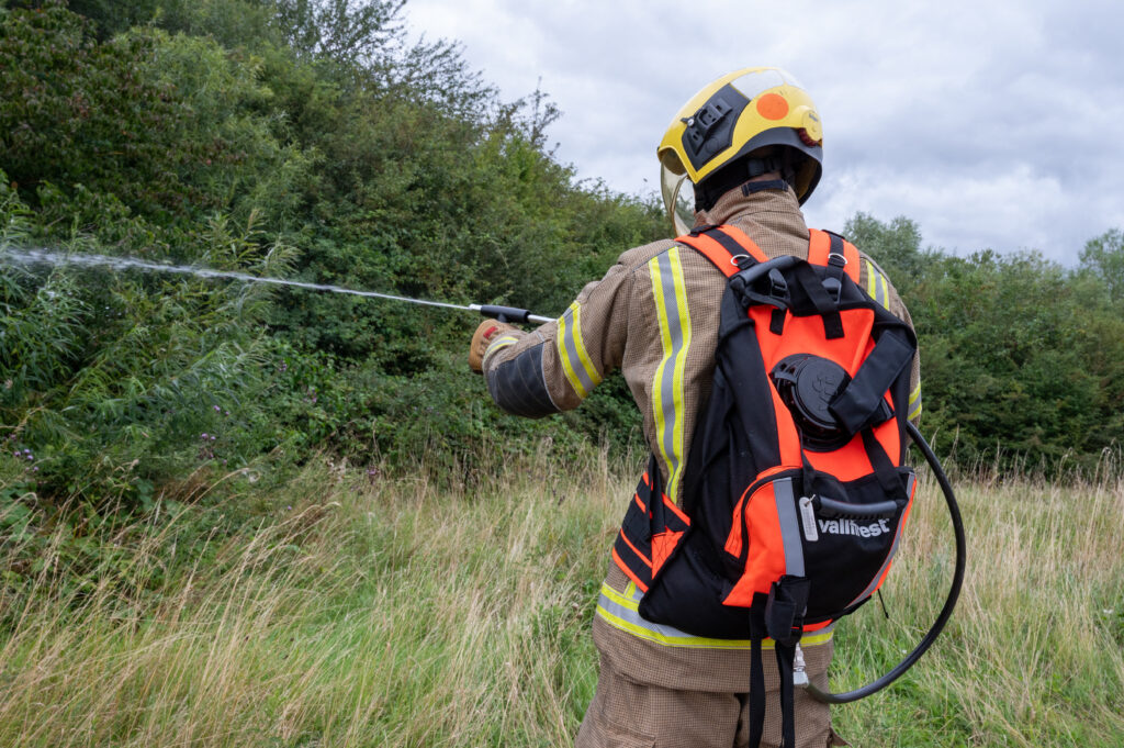 A firefighter wearing a fire tunic and yellow helmet is shown using the new wildfire backpack, which is orange and black, and is squirting water out