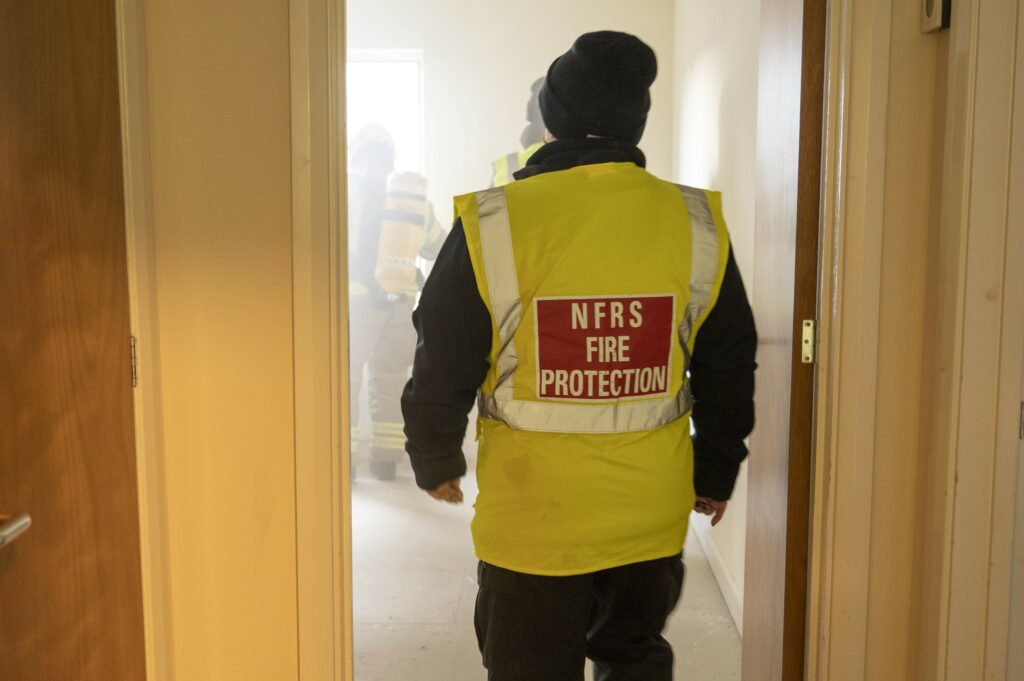 A Fire Protection Officer, wearing a fluorescent high-visibility jacket with the words NFRS Fire Protection on it, walks through a door into a smoke filled room