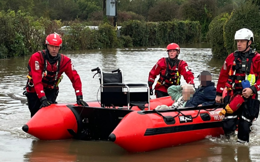 Three firefighters, wearing red helmets and uniforms, help to transport two people - whose faces are pixelated to protect their identity - from their flooded homes. A wheelchair is also on the boat. The flooding incident happened in another county.