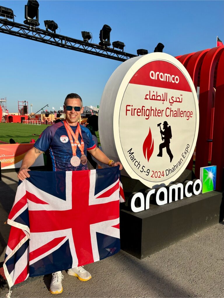 Dean Keeber, wearing a blue shirt with two red medals around his neck, poses with the United Kingdom flag next to a sign that says Aramco Firefighter Challenge, March 5-9, 2024 Dhahran Expo