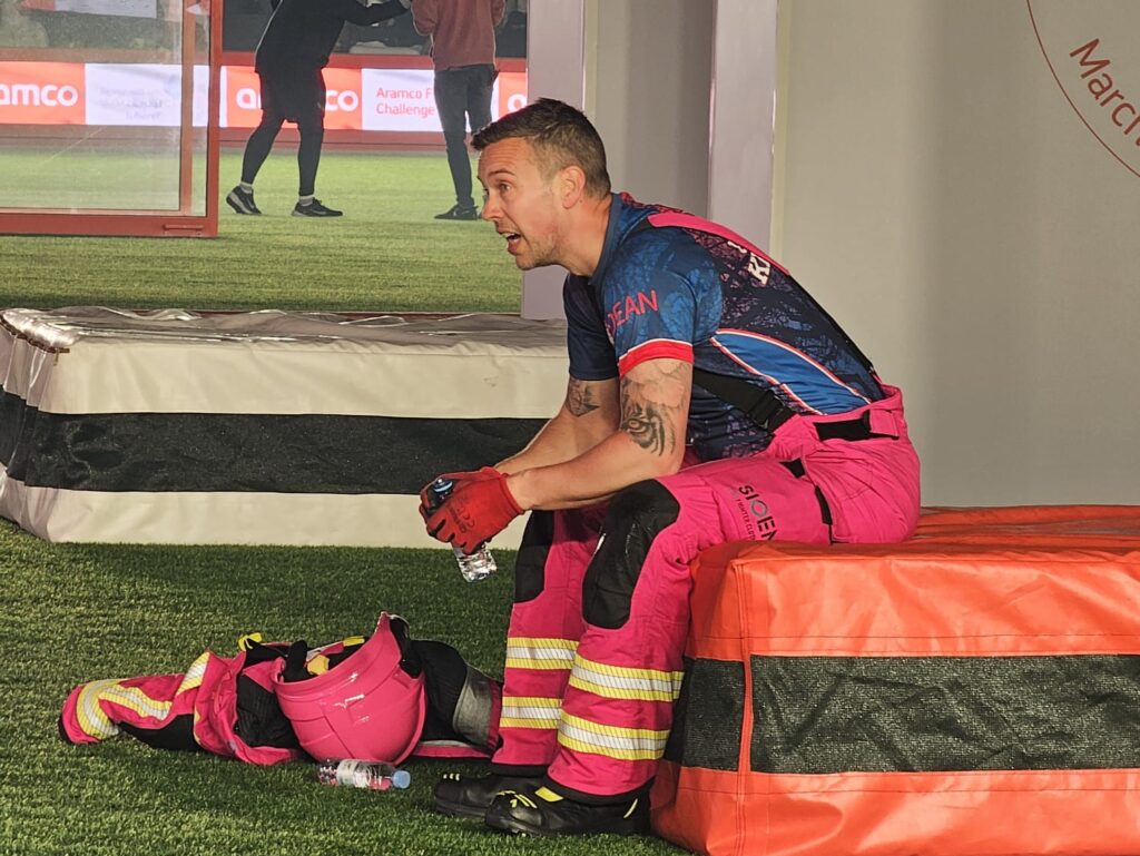 In the foreground is Dean Keeber, wearing a blue and pink fire tunic, who is show sitting down during a firefighter challenge.