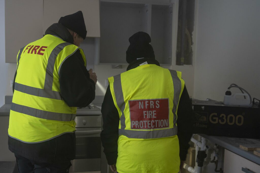 Two Fire Protection Officers for Northamptonshire, wearing fluorescent yellow jackets with the words 'NFRS Fire Protection' on them, examine a kitchen during an inspection.