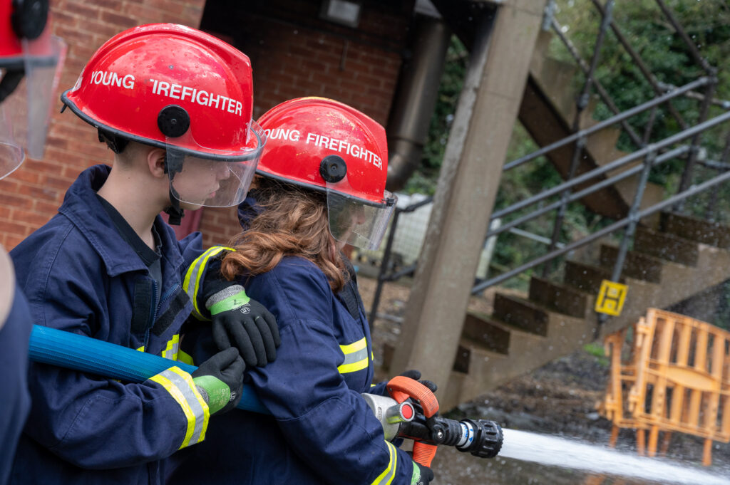 Two young people in fire kit squirting a hose
