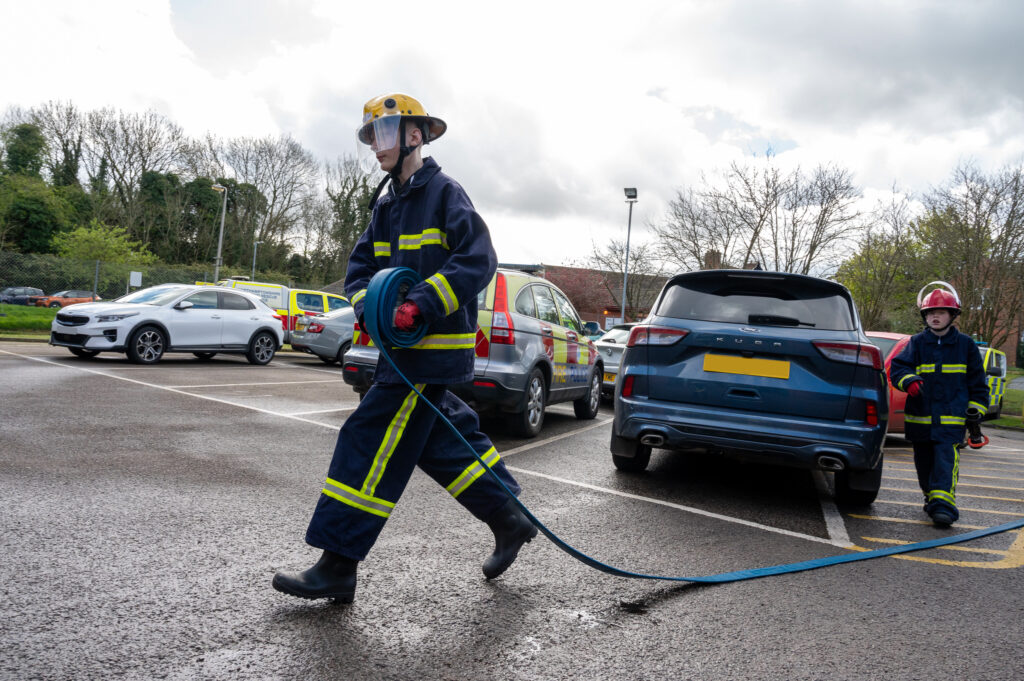 Two young people in fire kit, one carrying a hose