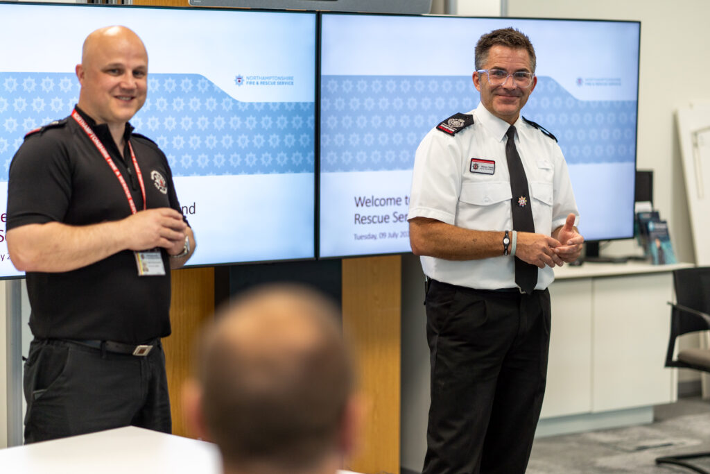 Pictured on the right, wearing a white shirt is Deputy Chief Fire Officer Simon Tuhill. Pictured on the left. wearing a black shirt, is Training Instructor Rich Wilding. The pair are pictured in front of a digital display showing the Northamptonshire Fire & Rescue Service branding.