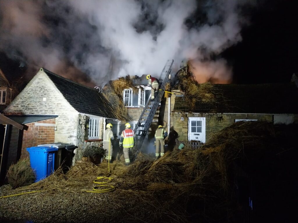 Four firefighters in full PPE and breathing apparatus use hoses and ladders to extinguish a fire in a thatched roof