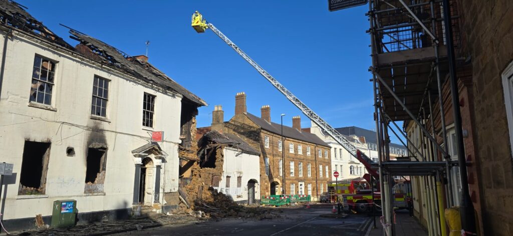 Street outdoors on a sunny day. Some building can be seen in the aftermath of a fire and one has partially collapsed. A fire aerial appliance can be seen with the ladder fully extended over the scene.