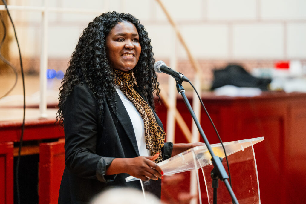Deputy PFCC Marianne Kimani wearing a black blazer and leopard scarf stands at a podium speaking into a microphone