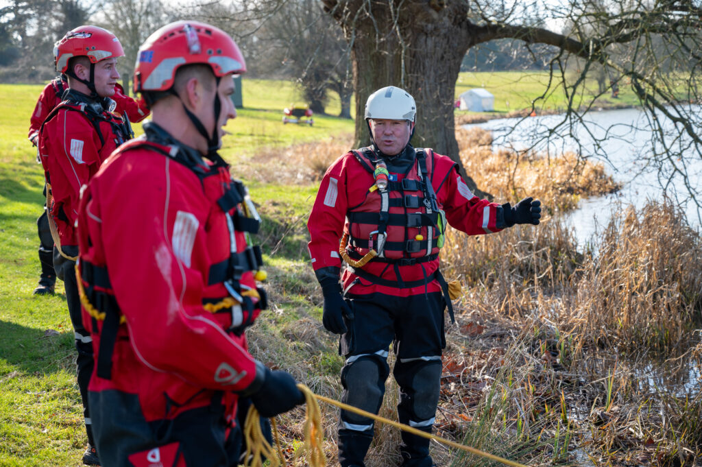 A training manager in full red water safety kit, teaching other firefighters on the water edge