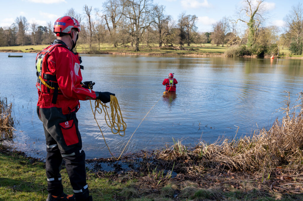 A firefighter, wearing red water rescue uniform, is shown holding a throwline to rescue another member of the Service, shown in the water also wearing red, during a water safety exercise.