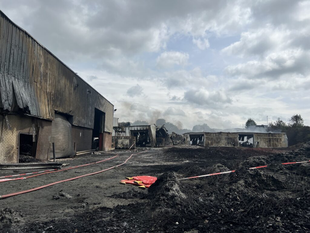 A large amount of burnt black plastic on the ground, with red fire hoses laid out in between it.
There is burnt and smoke damaged buildings in the background.