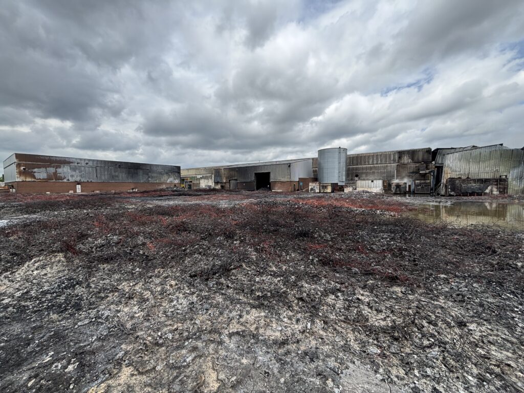 Photo shows a large amount of burnt plastic on the ground, with smoke damaged warehouse buildings in the background.