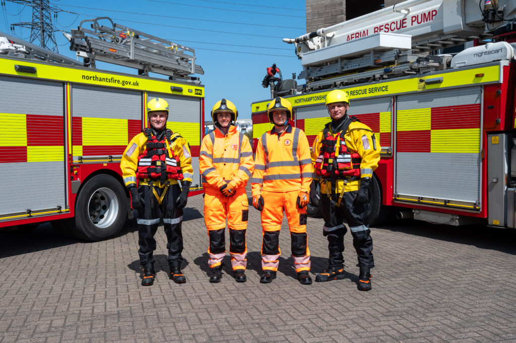 four people stand in front of two fire engines. Two people are wearing bright orange uniform. and two others are wearing black trousers with a yellow top and a red life jacket over the top.