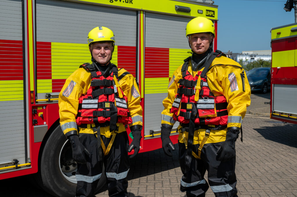 Two men stand in front of a red and yellow fire engine wearing black trousers, a yellow jacket, a red life jacket and yellow helmets