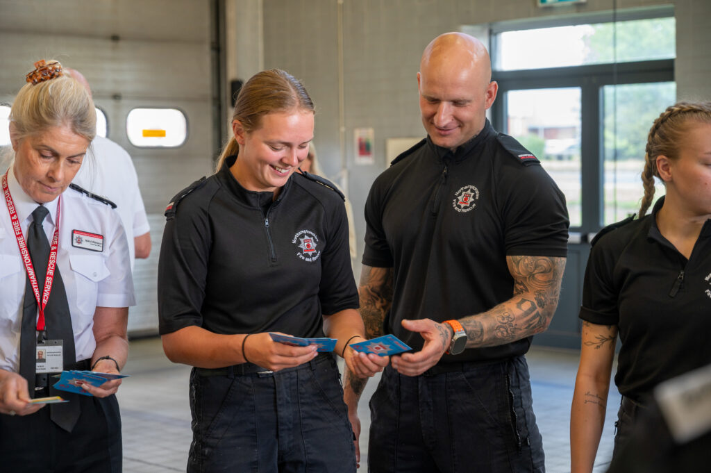 Four individuals in uniform stand indoors in a well-lit room with large windows and a garage door in the background. Three women and one man are visible; the two women on the left are holding blue cards and appear to be conversing with the man, who has tattoos on his arms. The woman on the far right is partially visible.