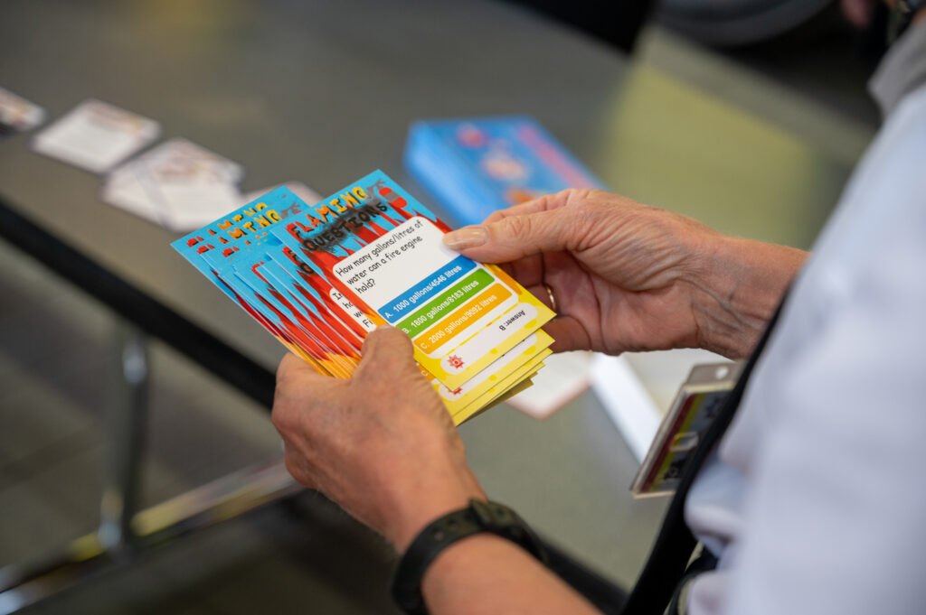a person holds a stack of colourful cards. The background is blurred, showing a table with more cards and papers scattered on it.