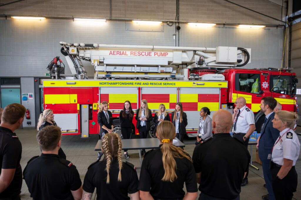 A group of people, including firefighters and civilians, are gathered inside a fire station. In the background is a large red and yellow fire truck labeled 'AERIAL RESCUE PUMP' and 'NORTHAMPTONSHIRE FIRE AND RESCUE SERVICE.' The group appears to be engaged in a discussion or presentation in front of the fire truck.