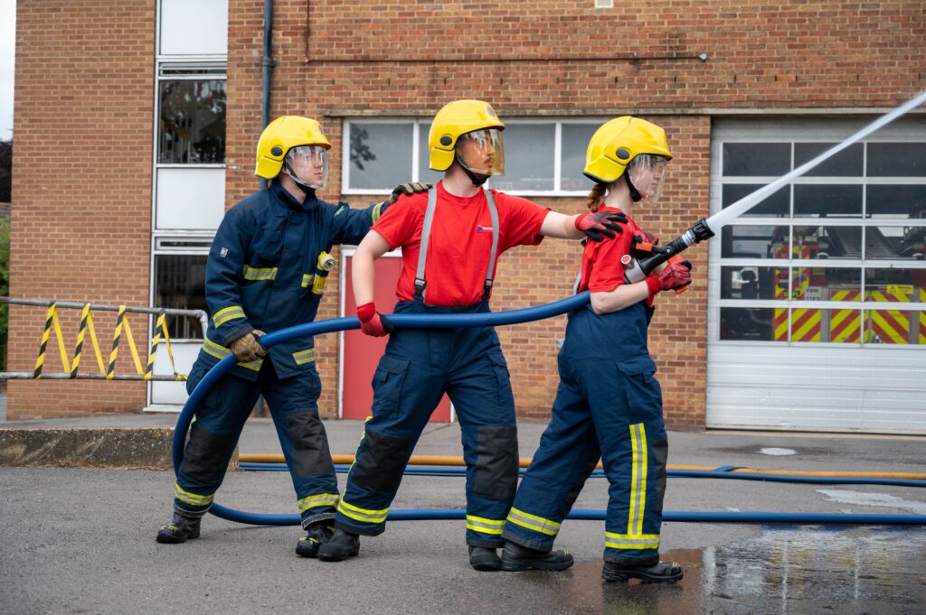 Three young people, wearing dark blue trousers and red t-shirts and yellow helmets hold a hose-reel jet pointing water upwards