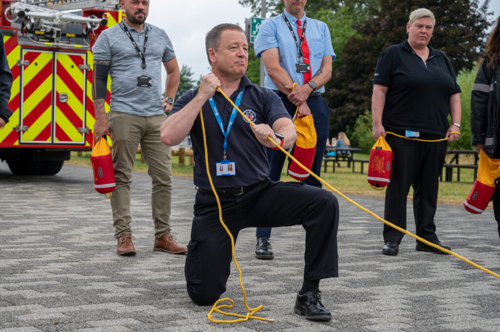 A man wearing a black t-shirt and black trousers and a blue lanyard throws a yellow throw line, behind him is two men and one woman standing.