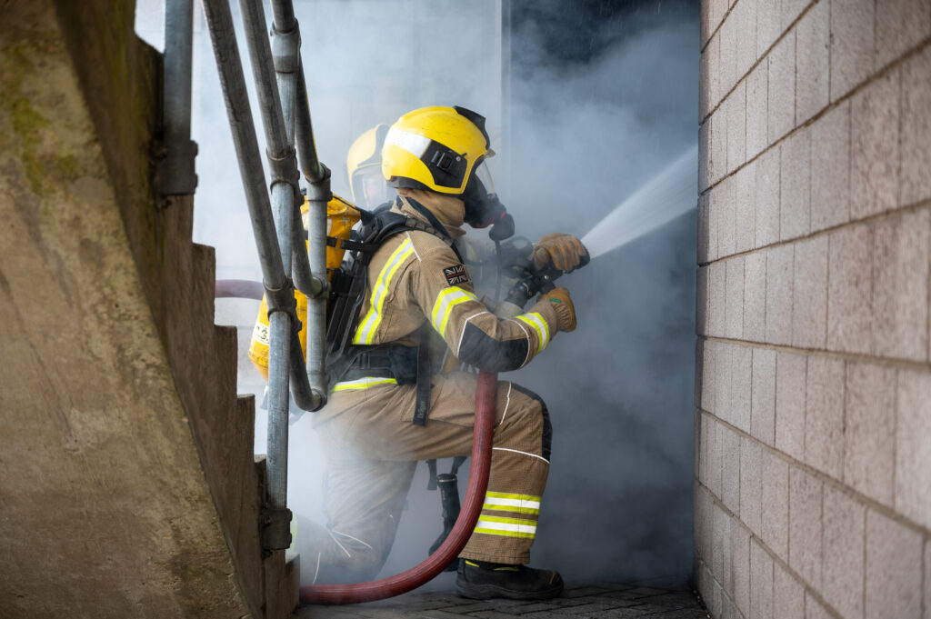 A firefighter in full fire kit squirting water in to a smoky building