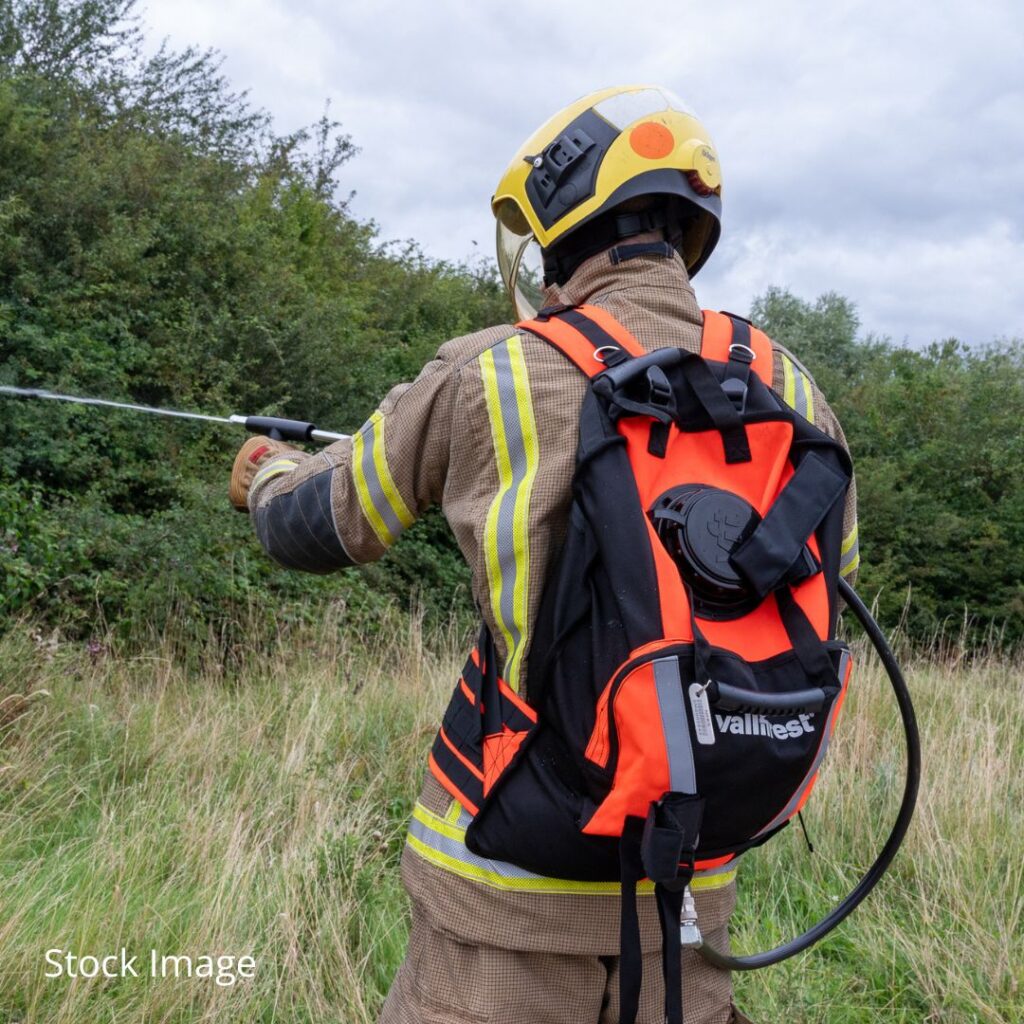 A firefighter in a yellow helmet and protective gear stands in a grassy area with dense green bushes, holding a hose or nozzle connected to an orange and black Vallfirest backpack