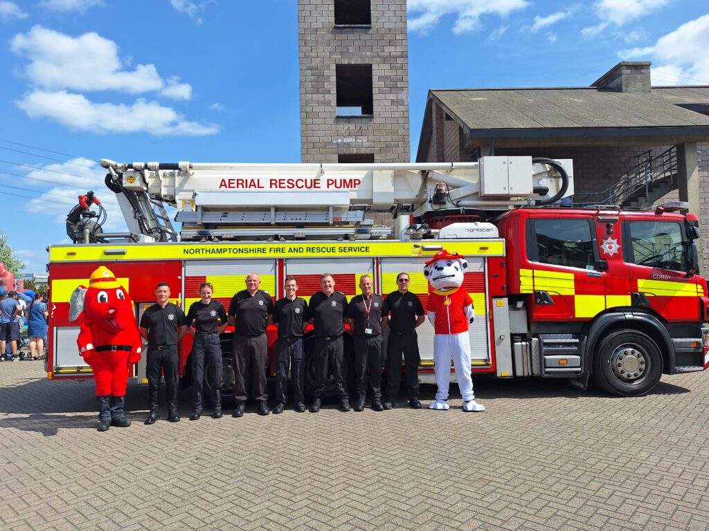 A group of firefighters stands in front of a large red fire truck labeled 'Aerial Rescue Pump' and 'Northamptonshire Fire and Rescue Service.' The firefighters are joined by two mascots, one dressed as a red elephant and the other as a white animal with a firefighter's hat. The background includes a tall brick training tower and another building. The sky is clear with some clouds.