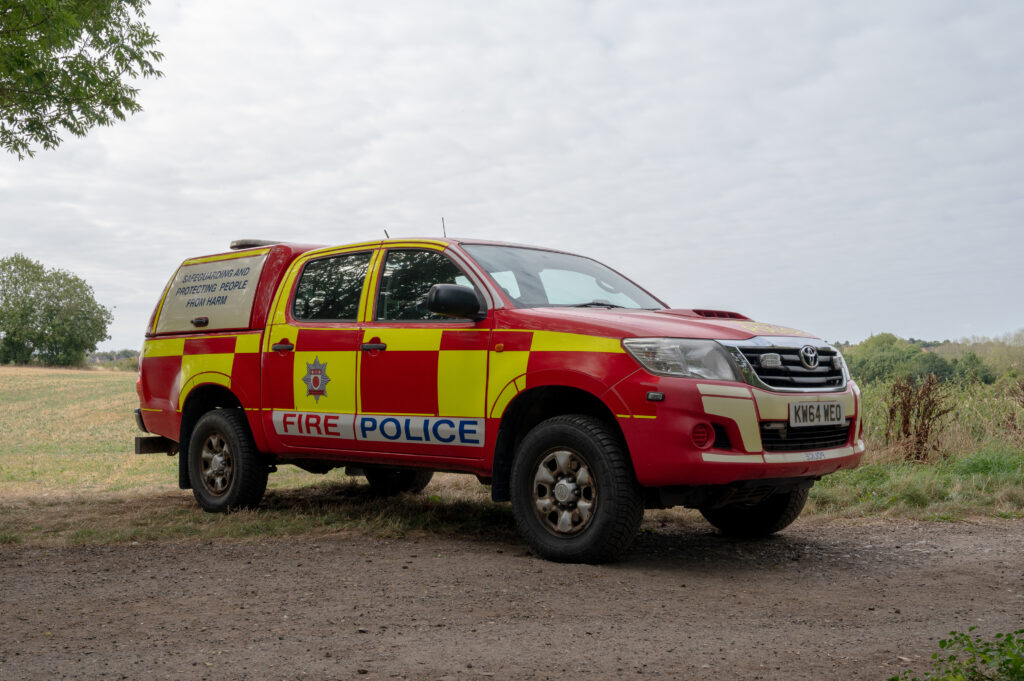 A red and yellow fire police vehicle is parked on a dirt path in a rural area with fields and trees in the background. The vehicle has 'FIRE POLICE' written on the side, along with an emblem. The license plate reads 'KN64 WEO.