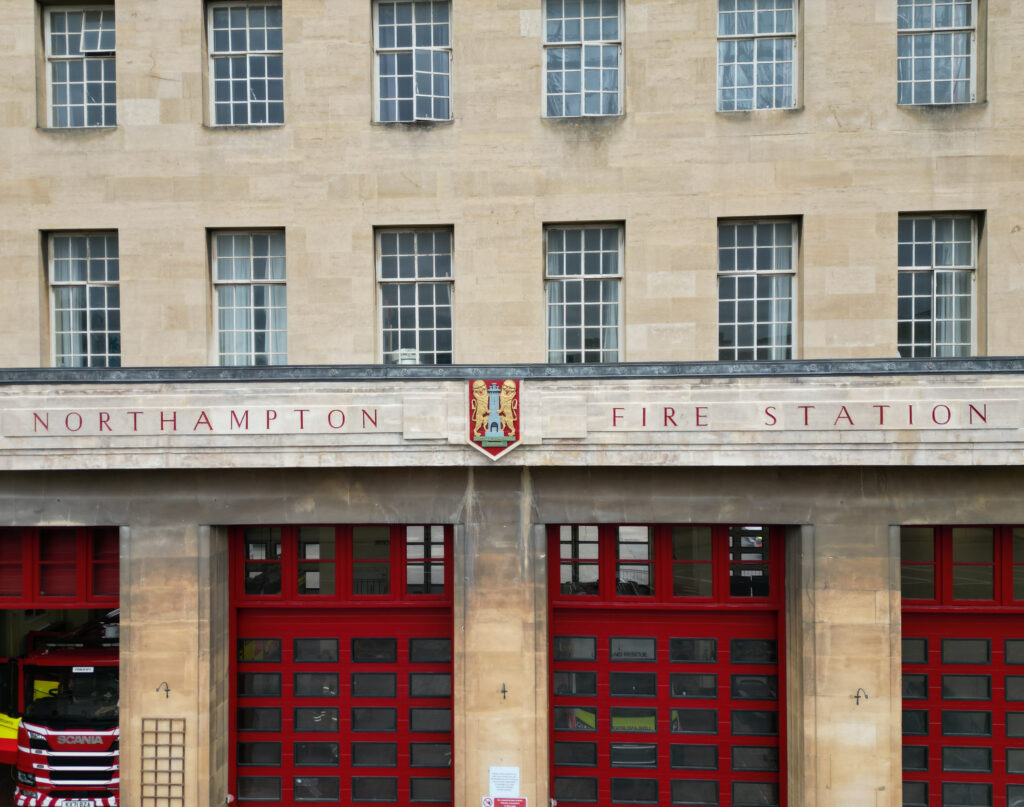 he image shows the front facade of the Northampton Fire Station. The building is made of beige stone with large rectangular windows on the upper floors. Below the windows, there is a sign that reads 'NORTHAMPTON FIRE STATION' in red letters, with a crest in the center. The ground floor features three large red garage doors, one of which is open, revealing part of a fire engine inside.