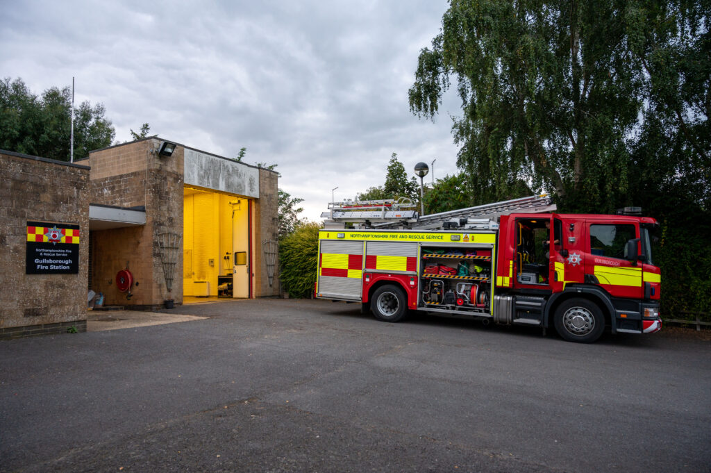 Fire Station with a red fire truck parked outside an open yellow garage door, surrounded by trees and bushes under a cloudy sky