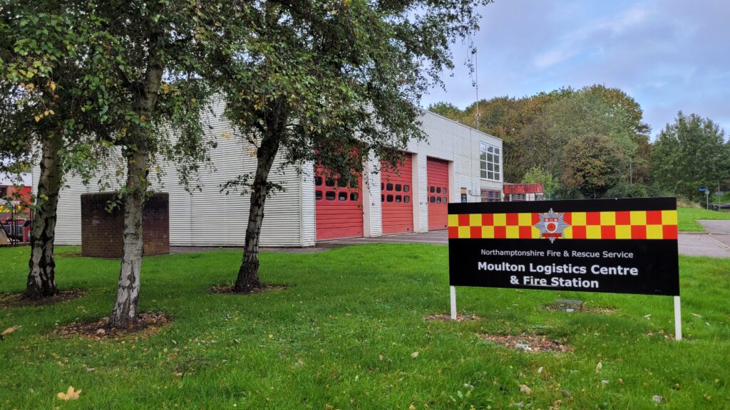 Exterior view of the Moulton Logistics Centre & Fire Station, part of Northamptonshire Fire & Rescue Service. The modern building features red garage doors and white siding, with a sign in front reading 'Northamptonshire Fire & Rescue Service Moulton Logistics Centre & Fire Station'. The surrounding area includes grass and trees.