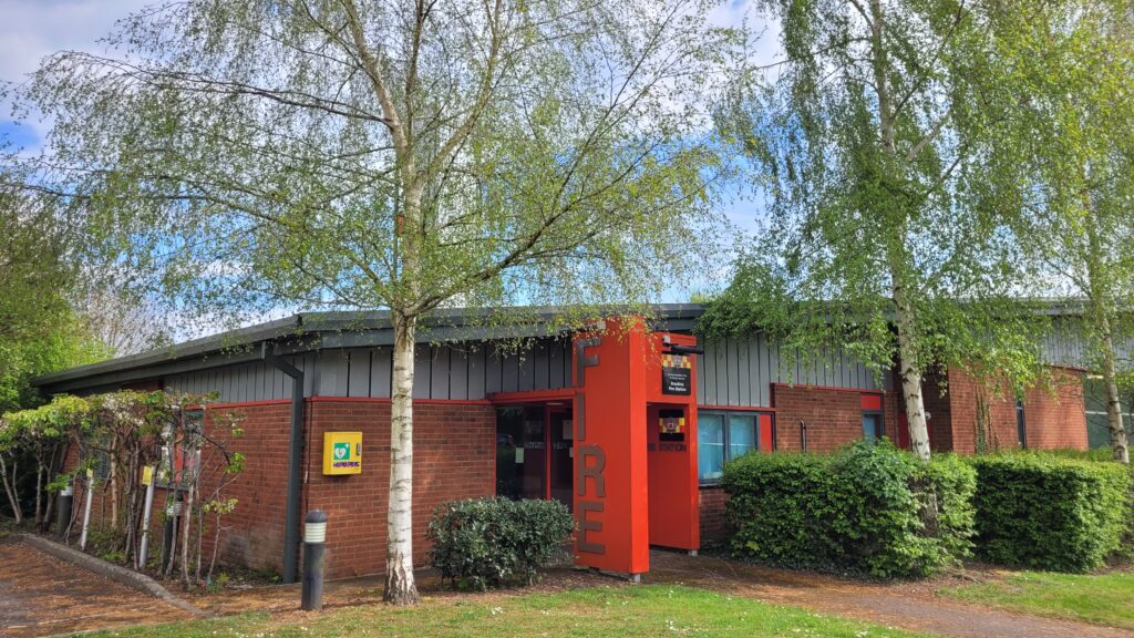 Brackley Fire Station with a red entrance labeled 'FIRE', brick exterior, surrounding greenery, and a yellow defibrillator box mounted beside the door
