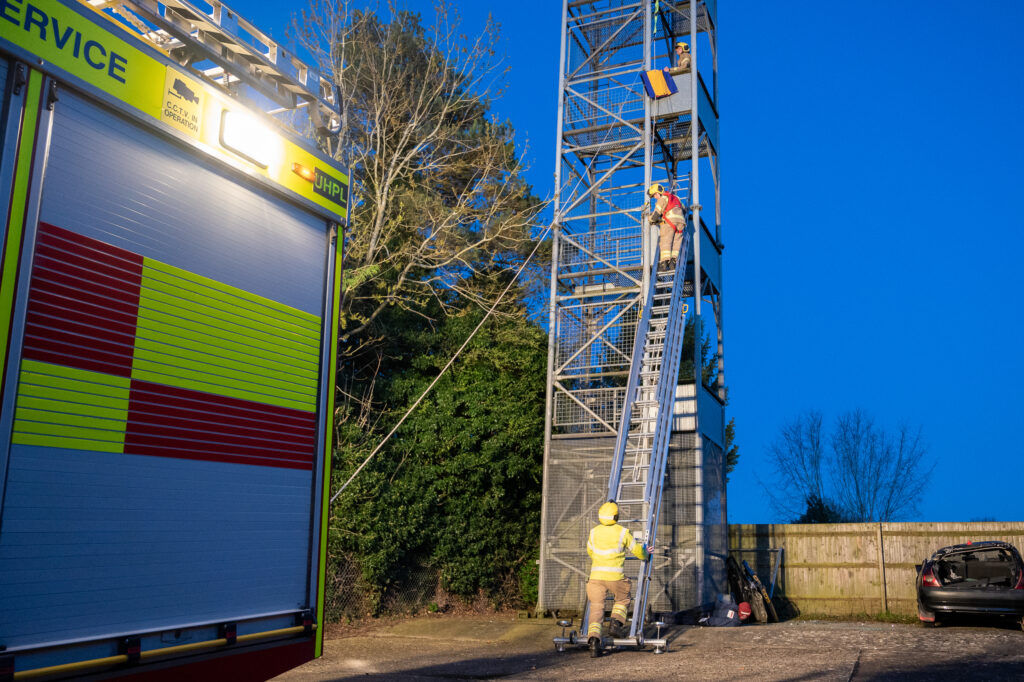 Firefighters in protective gear climbing a ladder against a tall metal structure during a training exercise, with a fire truck on the left and a damaged car near a fence in the background at dusk