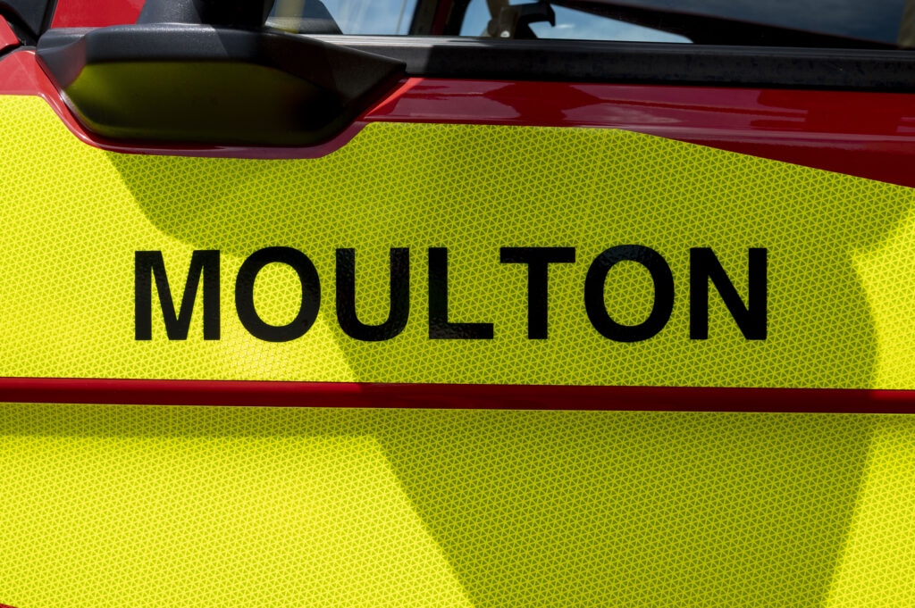 Close-up of a bright yellow reflective panel on a red emergency vehicle, featuring the word 'MOULTON' in bold black capital letters. The yellow surface has a textured pattern.