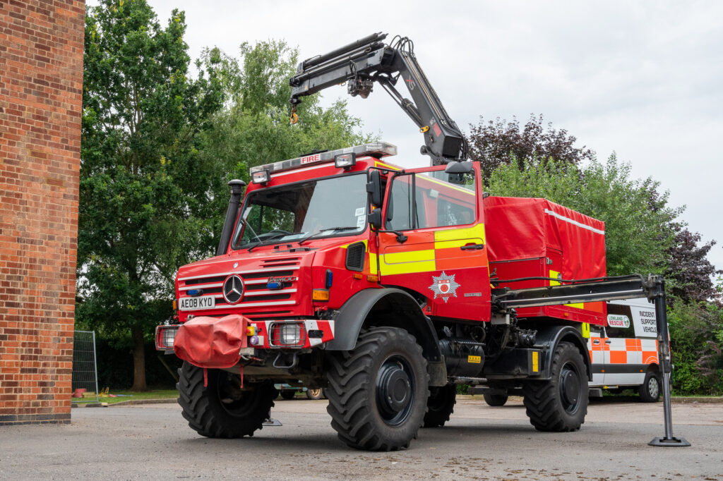 Red fire truck with a crane arm and large off-road tires, marked 'FIRE' on the front and bearing the license plate 'AE08 KYO'. An emblem on the side indicates it belongs to a fire and rescue service. In the background, an 'Incident Support Vehicle' is parked near trees and a brick building.