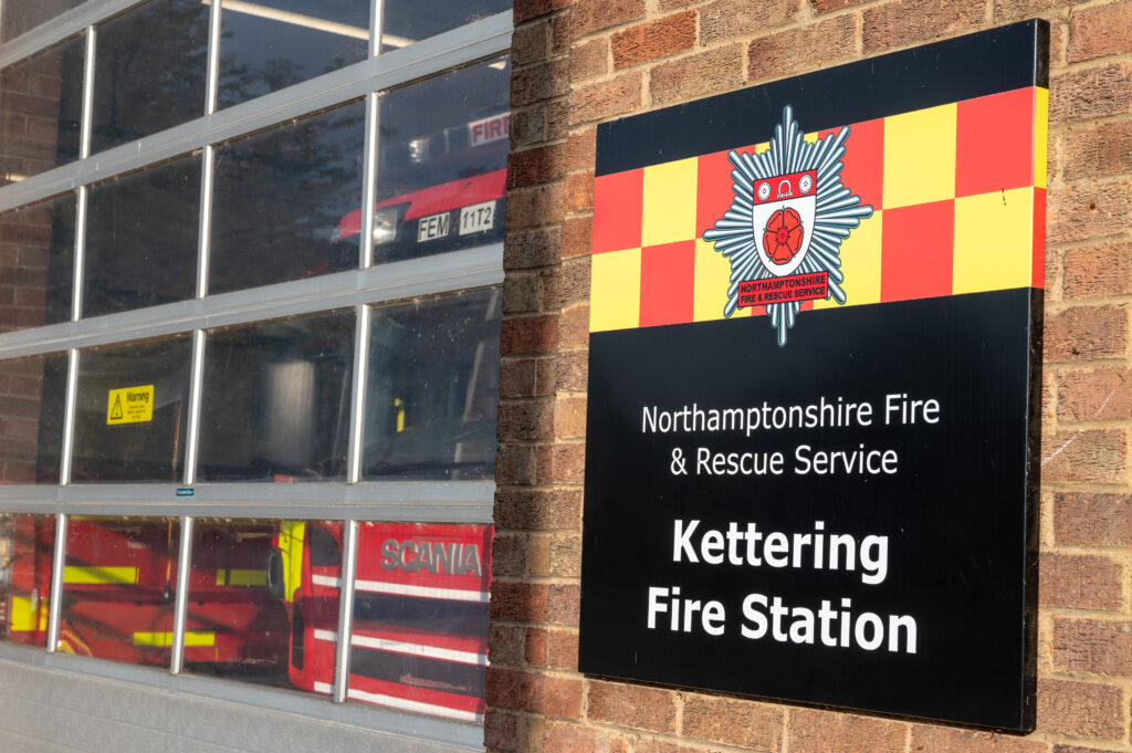 Exterior of Kettering Fire Station, part of Northamptonshire Fire & Rescue Service. A black sign with white text displays the station's name and emblem featuring a red rose and crossed axes on a yellow and red checkered background. A large glass garage door reveals parts of fire trucks inside, including one marked 'Scania'."
