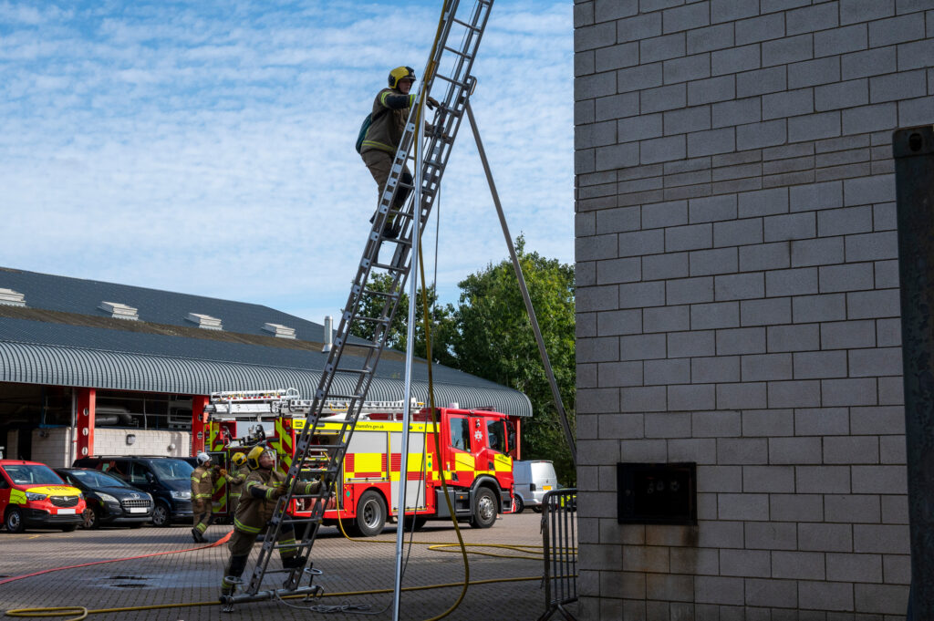 Firefighters performing a ladder drill outside a fire station. One firefighter climbs a tall, angled ladder while two others stabilize it at the base. A red and yellow fire truck and other vehicles are parked nearby under a partly cloudy sky, with trees and a brick building in the background.