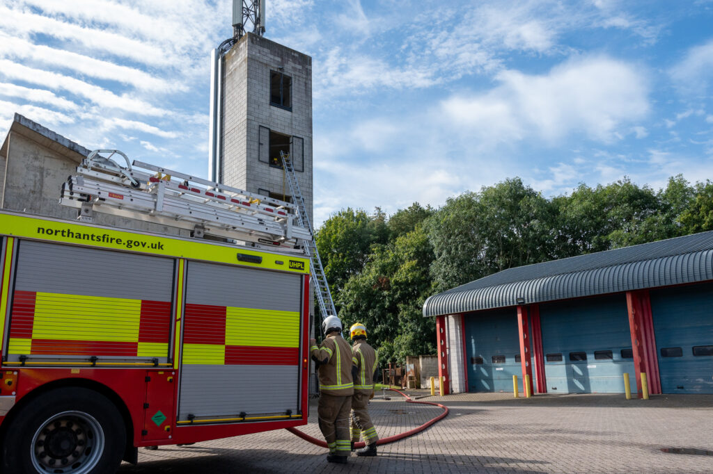 Two firefighters in full gear stand beside a fire truck marked 'northantsfire.gov.uk'. A ladder extends from the truck to a tall training tower, where another firefighter is visible through an open window. The scene is set in an open area with a building featuring multiple garage doors, surrounded by trees under a partly cloudy sky.